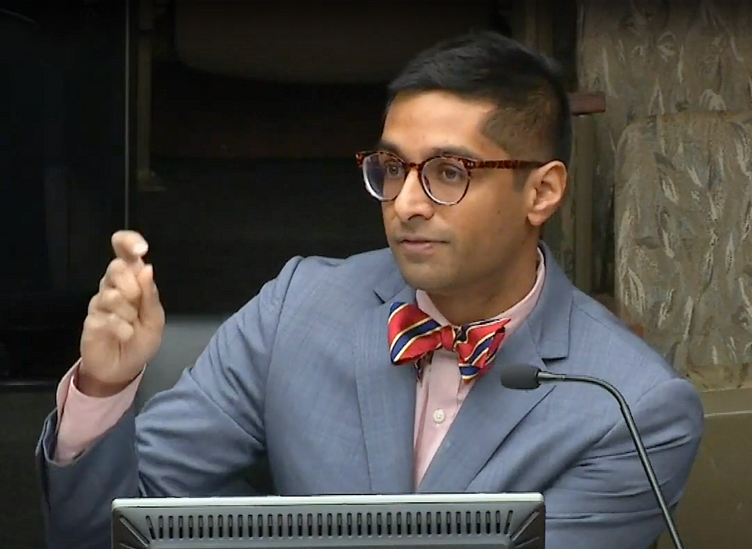A man wearing glasses, a light blue suit, a pink shirt, and a red, blue, and yellow striped bow tie seated at a table with a microphone and a computer monitor in front of him, gesturing with his hand.