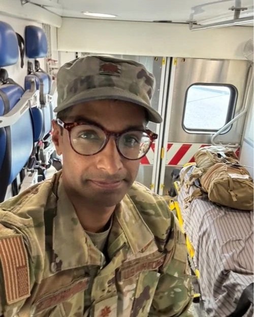 A soldier wearing camouflage uniform and cap, with glasses, sitting inside a military aircraft with seats and a bed in the background, partially covered with a bedsheet and other items.
