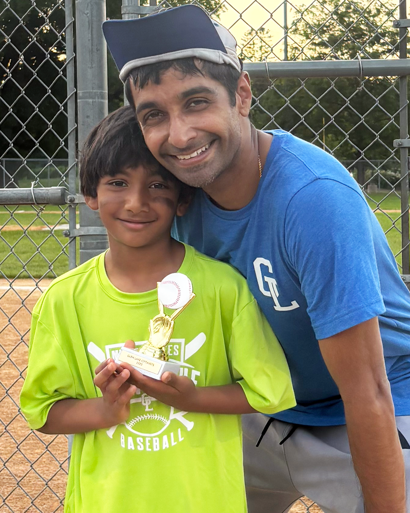 A young boy holding a small baseball trophy and a baseball, smiling, standing next to an adult man in a blue shirt, both outdoors on a baseball field with a chain-link fence and trees in the background, during sunset.