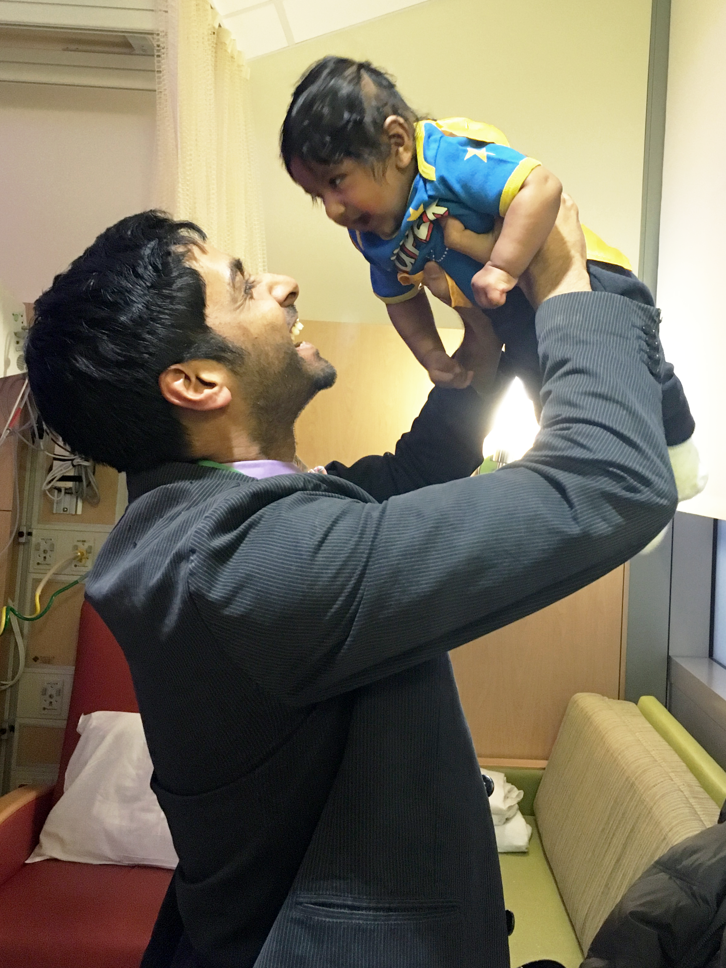 A man lifting a young boy in the air in a hospital room, both smiling and looking at each other.