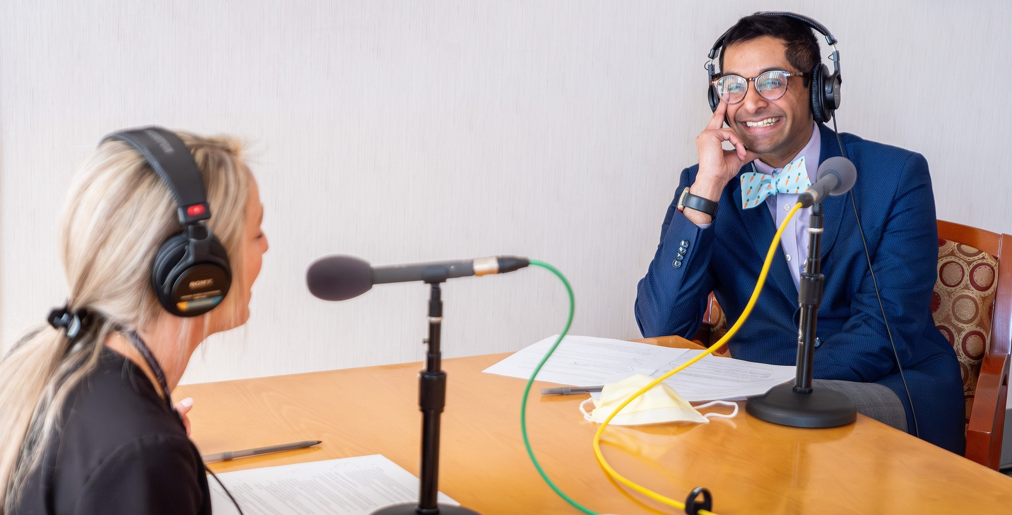 Two people, a woman and a man, are sitting at a table with microphones, headphones, and papers, engaged in a lively conversation or recording session; the man is smiling and wearing a blue suit and bow tie.