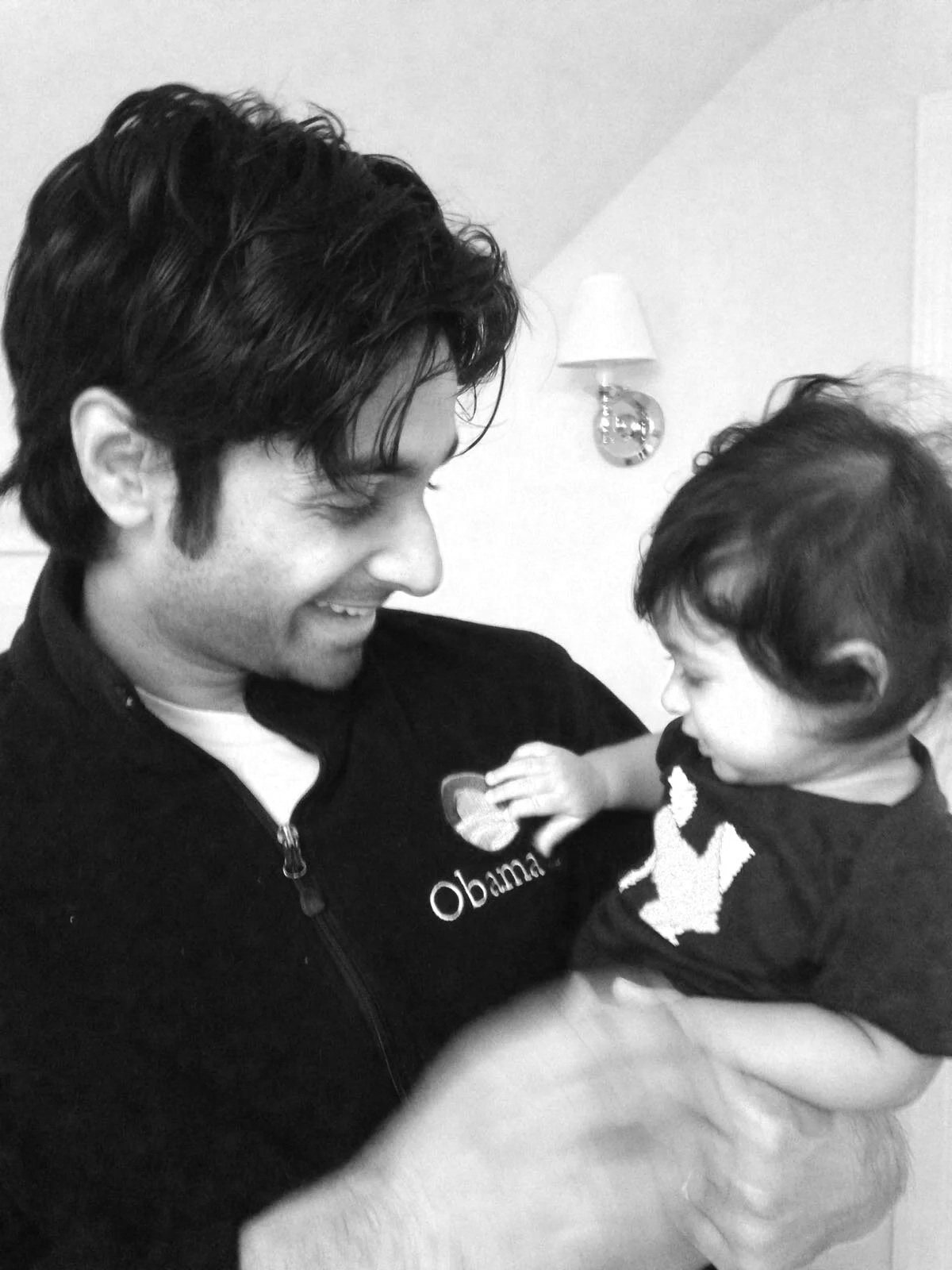 Black and white photo of a man with dark hair smiling while holding a small child, who is touching a button on his shirt. The man is wearing a jacket with the word 'Obama' and an emblem on it.