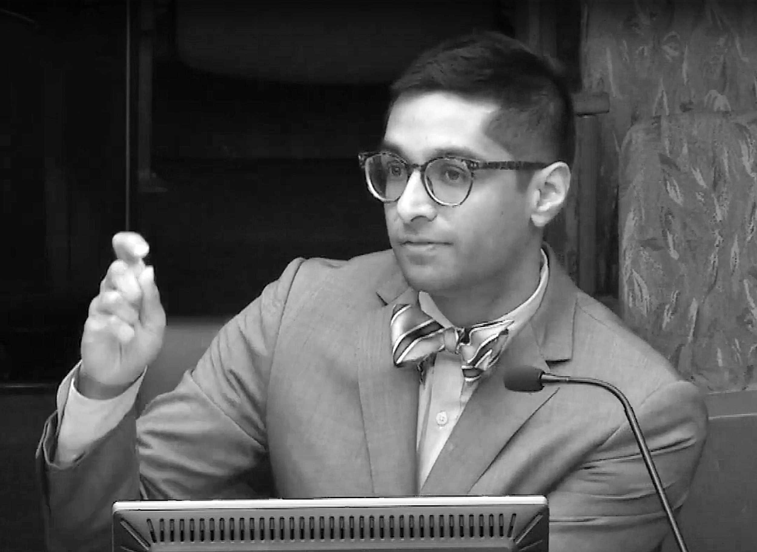 Black and white photo of a man wearing glasses, a suit, and a striped bow tie, seated at a table with a microphone in front of him, gesturing with his right hand.