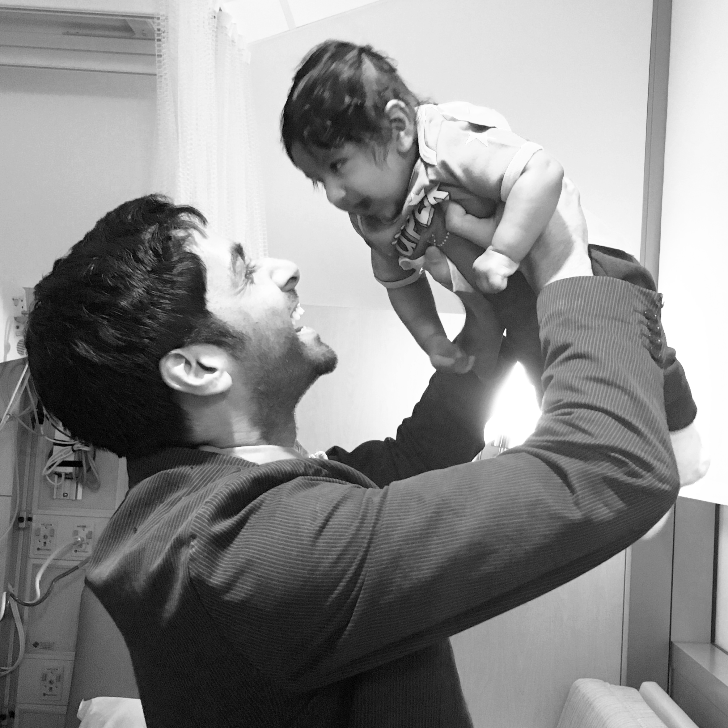 A man with dark hair and a beard holding up a young child in a hospital room, both smiling and looking at each other.
