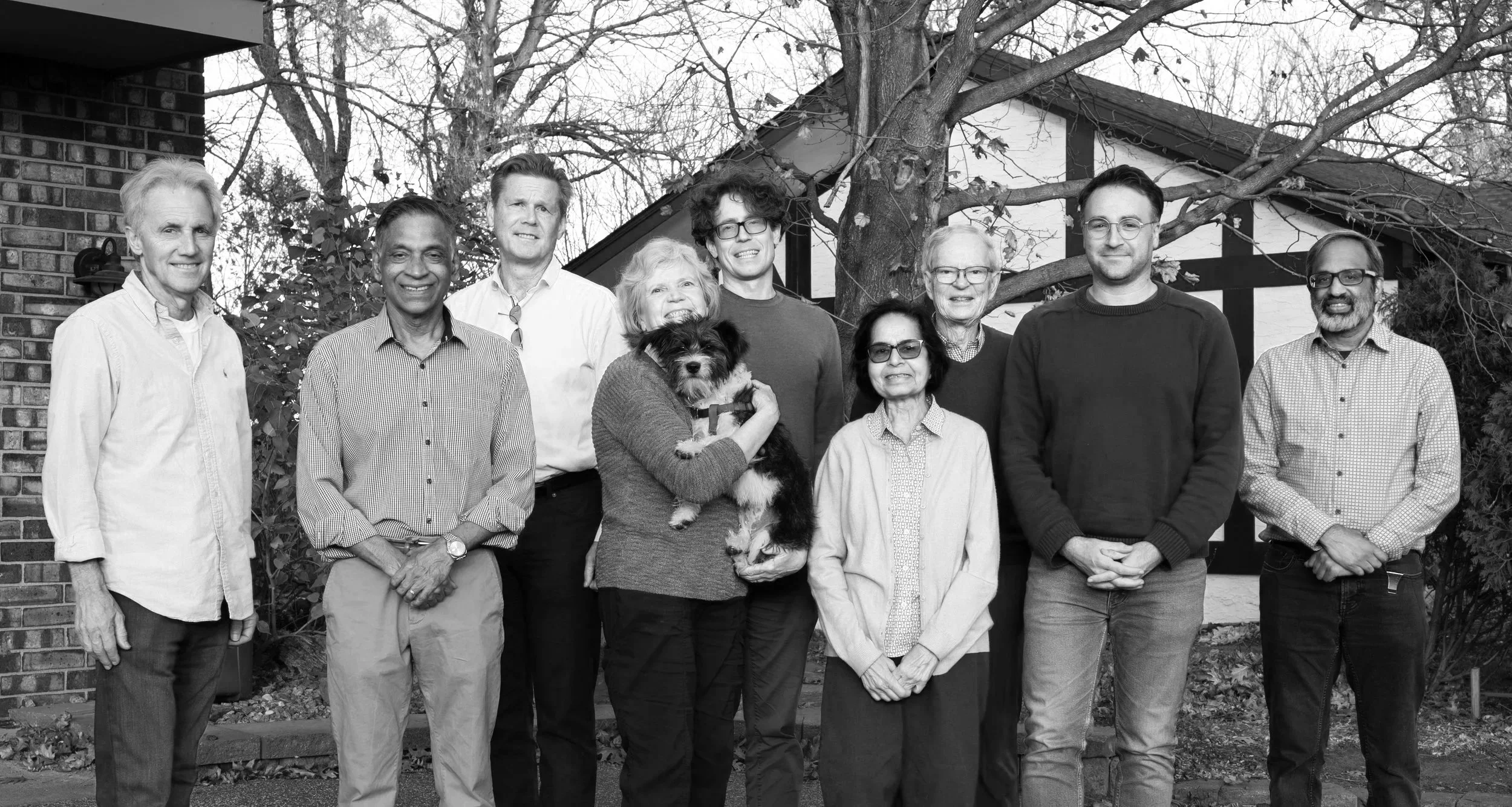 A group of ten people and one dog standing outdoors in front of a house with trees in the background, black and white photograph.