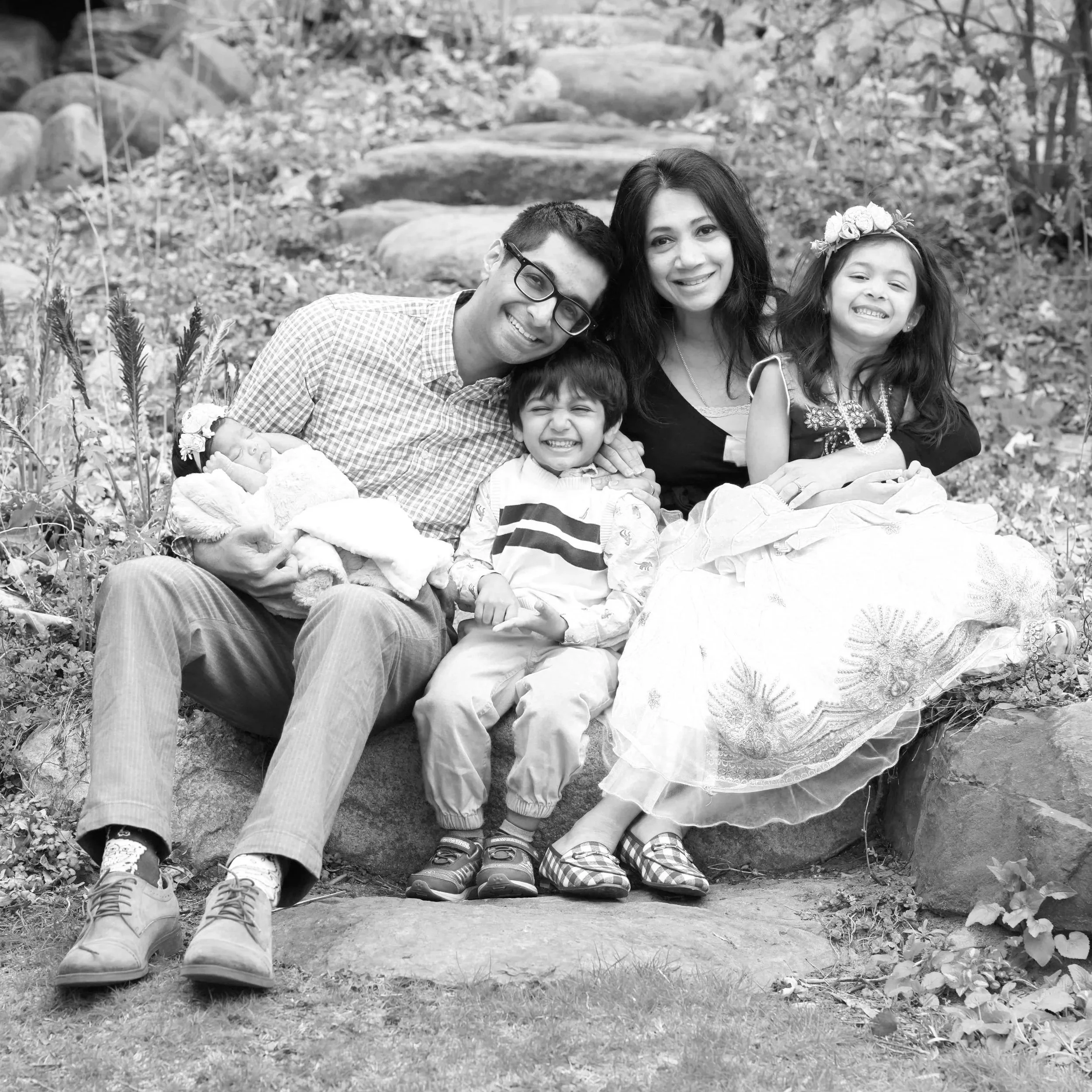 A family sitting outdoors on rocks in a natural setting. They've gathered for a photo, smiling, with a baby in the arms of a man on the left, two young children, and a woman sitting on the right. The background is filled with rocks and foliage.