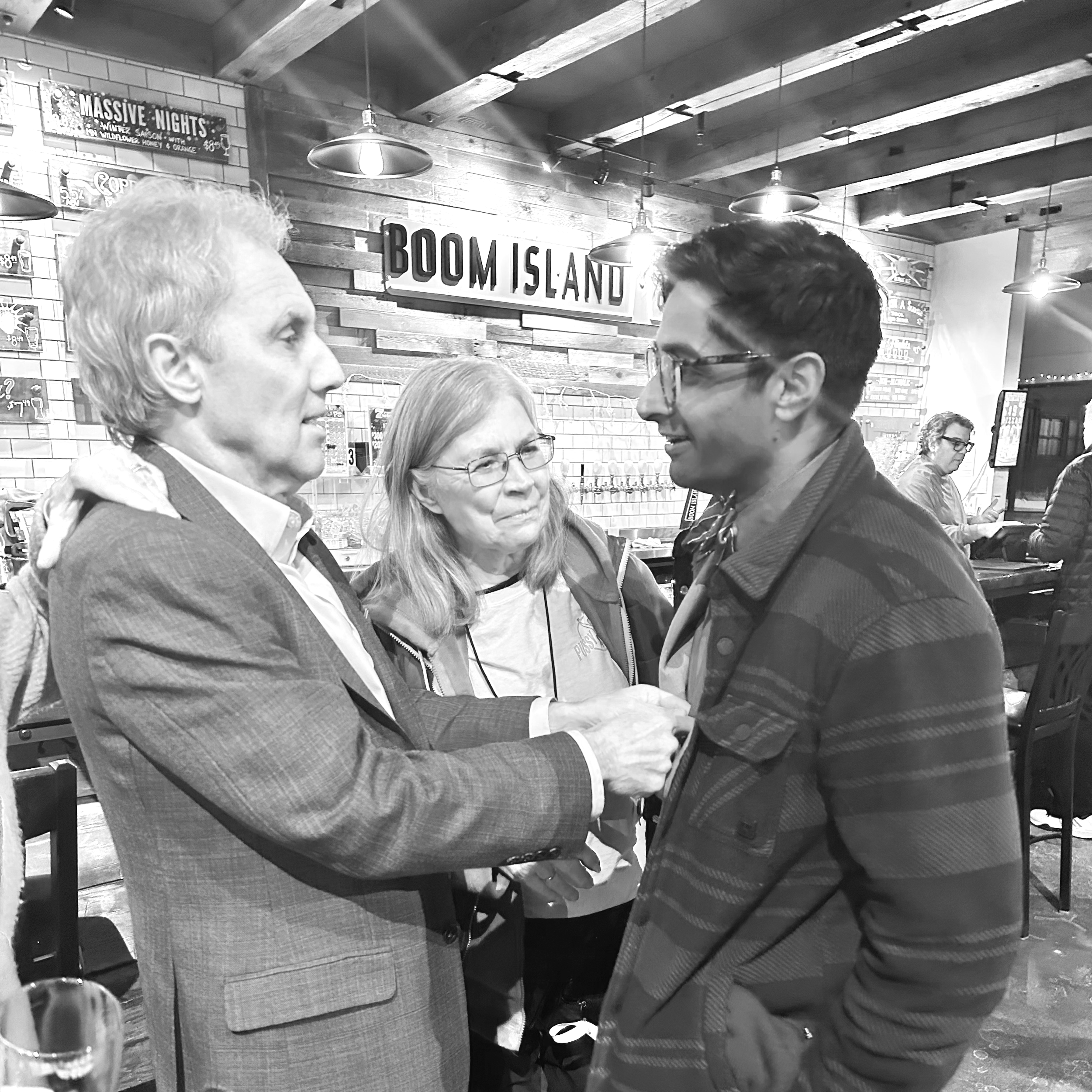 Three people talking in a bar with a sign that says 'BOOM ISLAND' in the background.