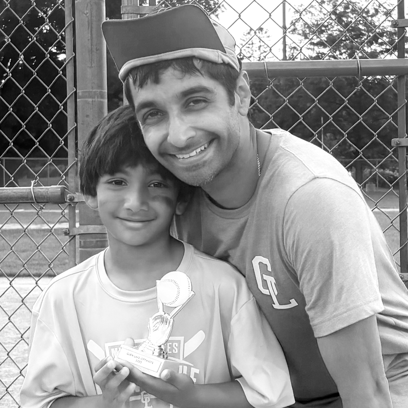 A young boy holding a trophy and an adult man smiling next to him, both in sports attire, at a baseball field with a chain-link fence in the background.