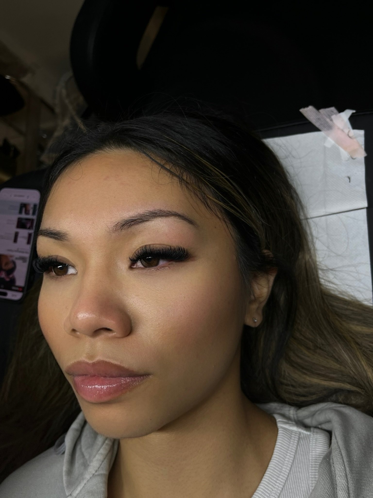 Close-up of a woman with makeup, long eyelashes, and earrings, lying down. There is a smartphone nearby showing a social media profile.