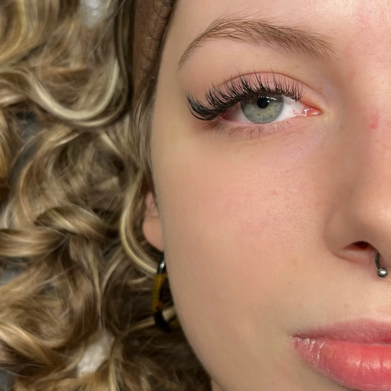 Close-up of a woman's face focusing on her right eye, with blonde curly hair, light skin, and a septum piercing.