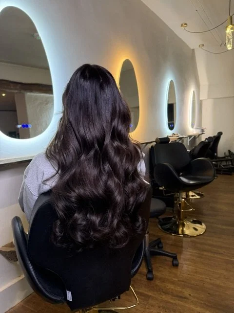 Back of a woman with long, wavy, dark hair sitting in a salon chair inside a modern hair salon with white curved walls and round mirrors.