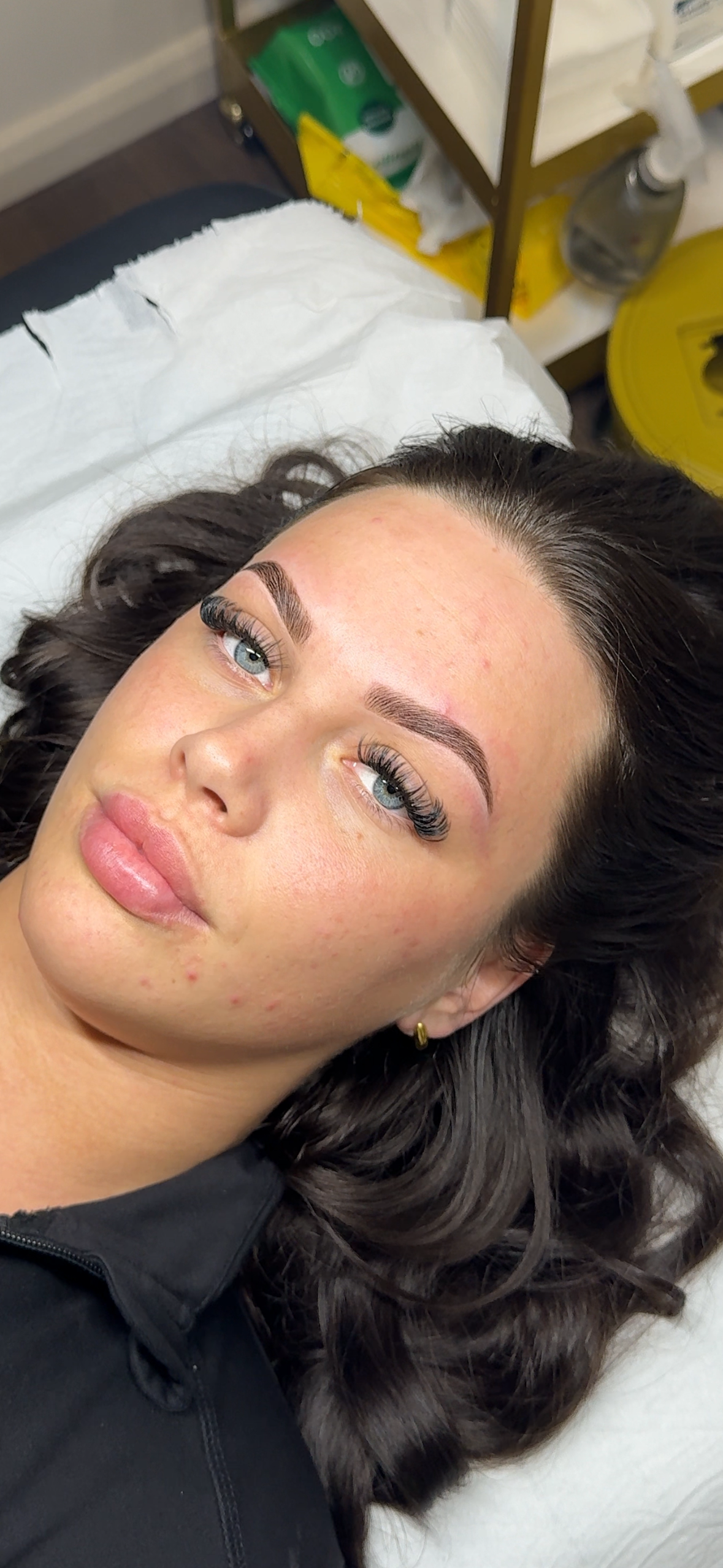 A woman lying on a bed with freshly done eyelash extensions and well-groomed eyebrows, with a background showing a shelf with cleaning supplies.