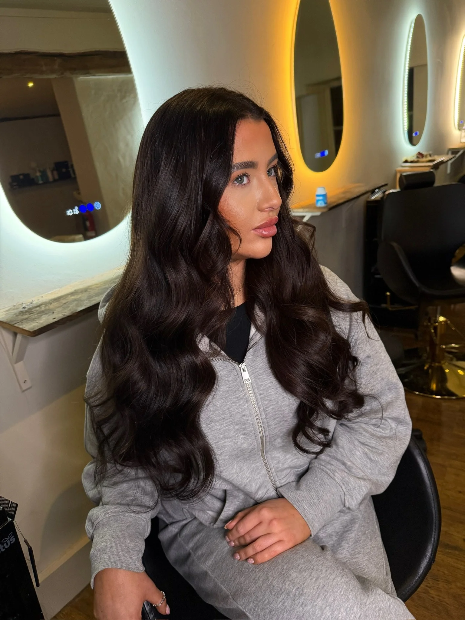 A woman with long, dark, wavy hair sitting in a salon chair in front of illuminated mirrors.