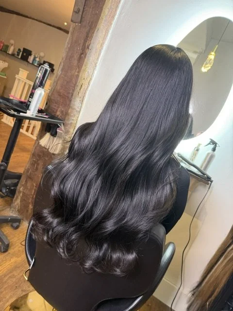 Back view of a woman with long, shiny, wavy black hair in a salon chair.
