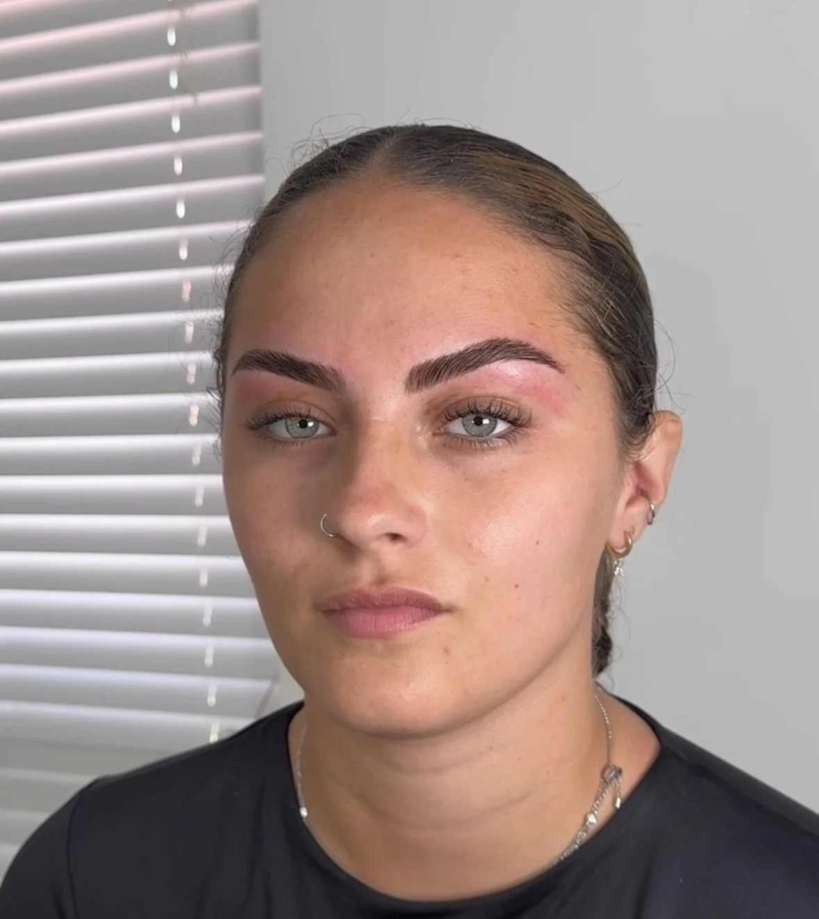 Close-up of a woman's face with light makeup, multiple earrings, and a nose ring, standing in front of a window with white blinds.