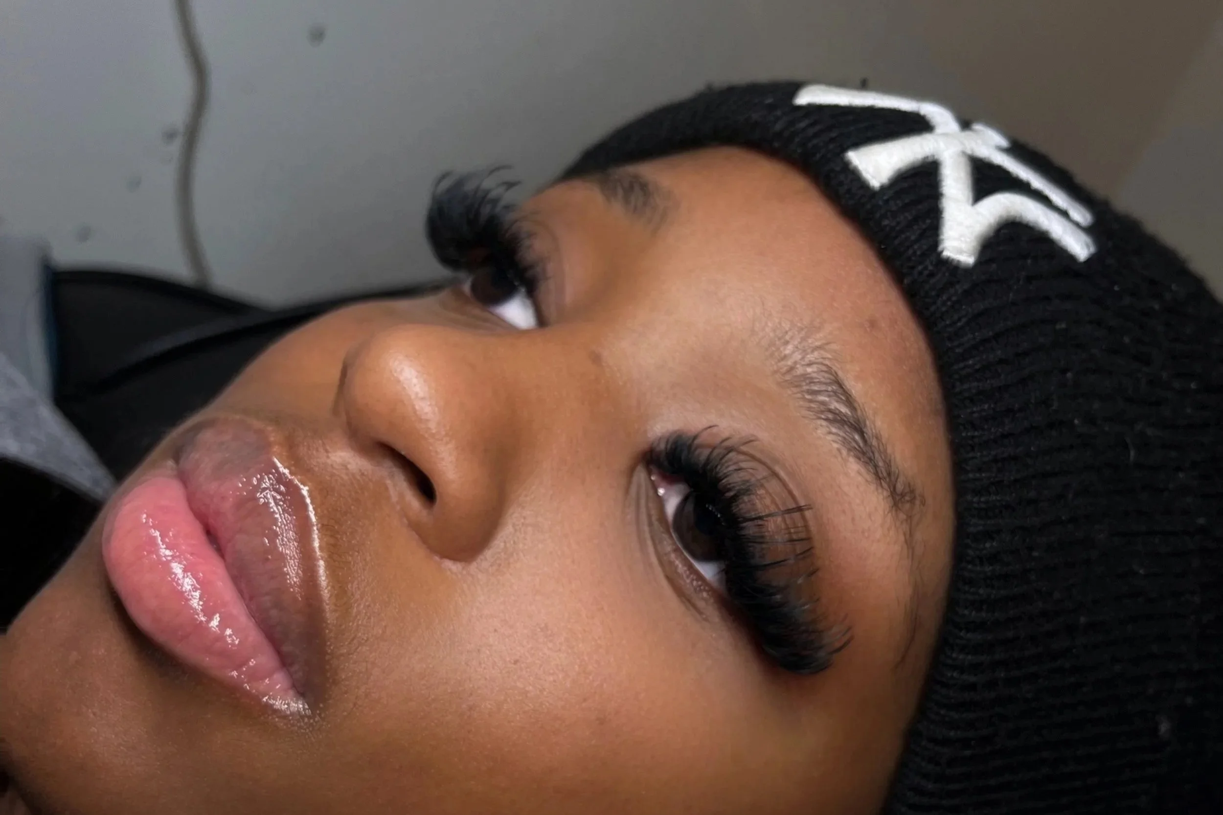Close-up of a woman's face lying down, wearing makeup and false eyelashes, with a black knit beanie with white lettering.
