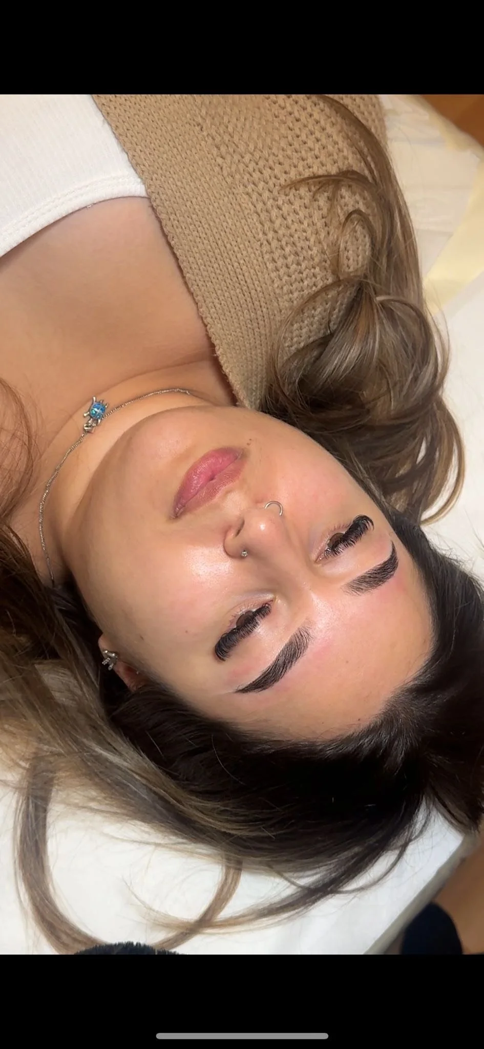 Woman with dark hair and multiple earrings lying on a bed, wearing a beige and white top, looking at the camera.