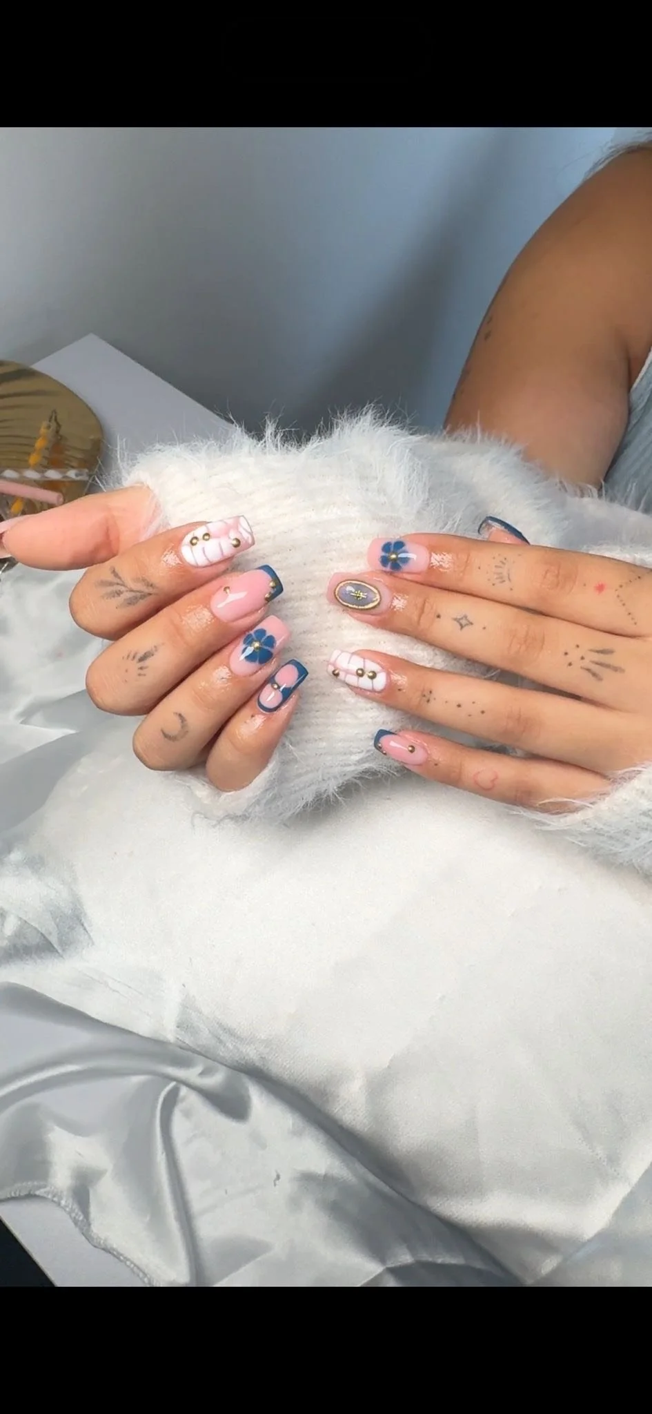 Person showing hands with decorated manicure, featuring blue flowers, gold accents, and a French tip style, on a white fluffy surface.