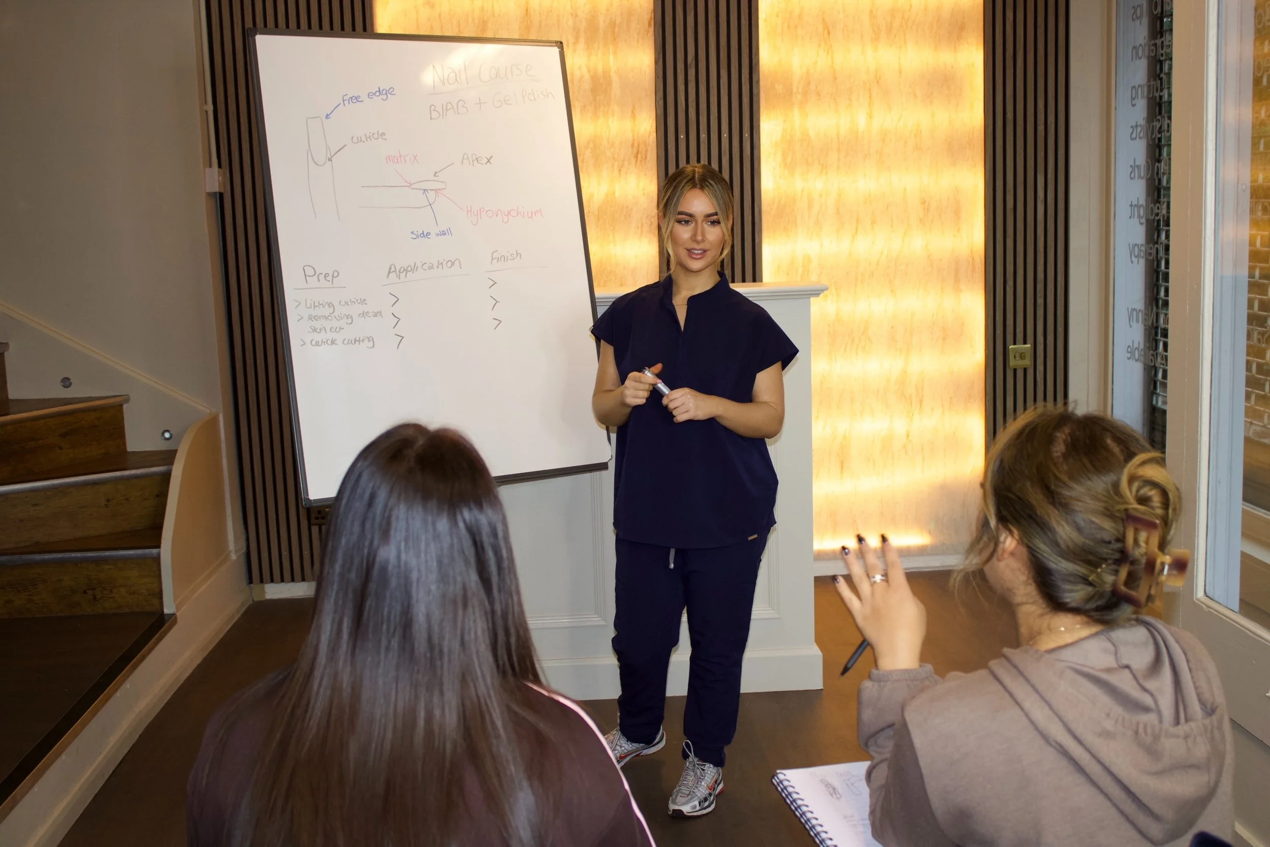 A woman standing in front of a whiteboard giving a presentation to two seated women in a room with wooden stairs, striped accents, and warm lighting.