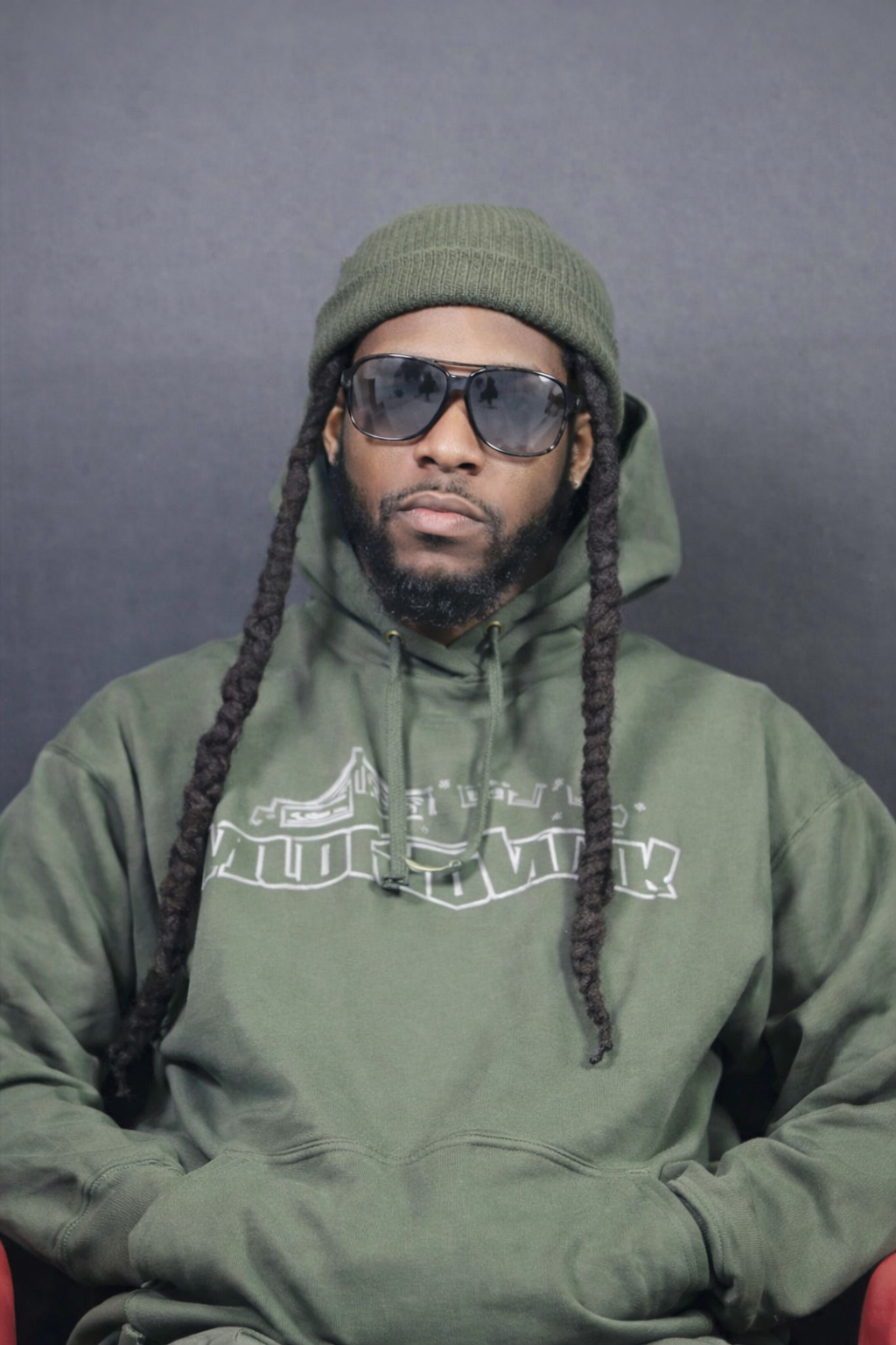 A man with long dreadlocks, wearing a green beanie, sunglasses, and a green hoodie, sitting against a plain dark background.