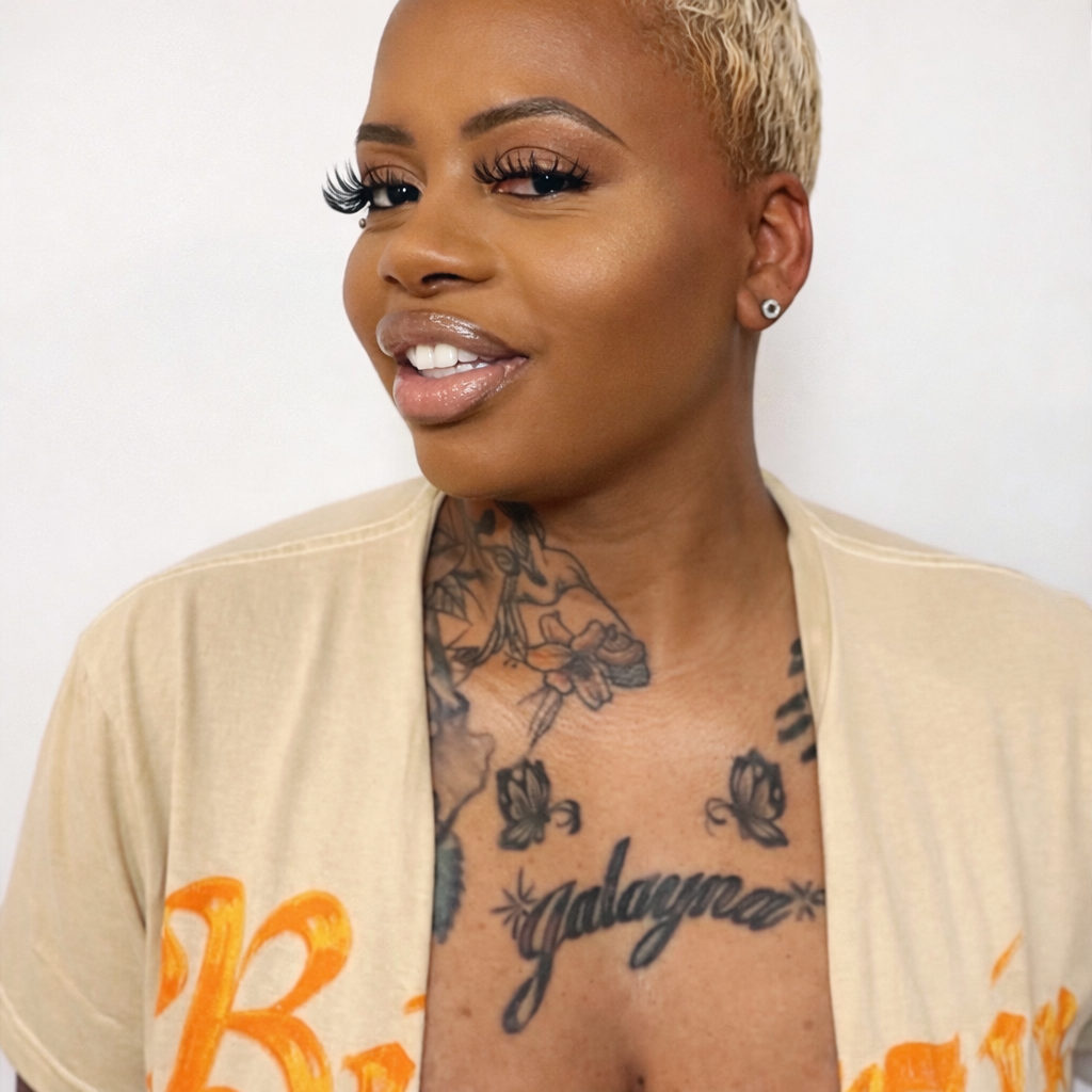 A woman with short blonde hair, makeup, and tattoos on her chest and neck, winking and smiling while wearing a beige shirt with orange writing.