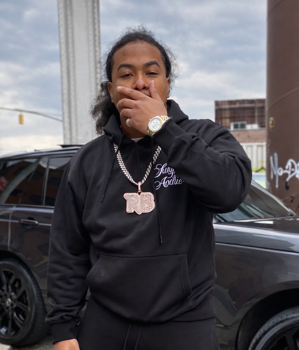 Young man wearing black hoodie with 'Stay Academy' written on it, jewelry, and a watch, posing with hand covering mouth in an urban setting with cars and buildings in the background.