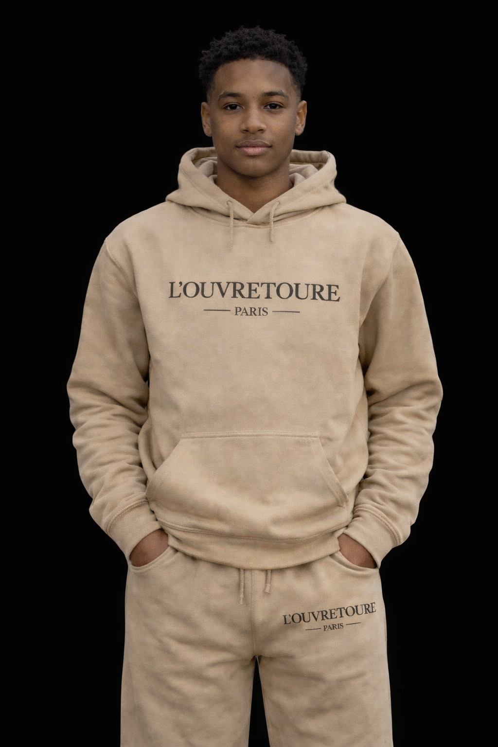 A young man wearing a beige hoodie and sweatpants with 'L'OUVRETOURE PARIS' written on them, standing against a black background with his hands in his pockets.