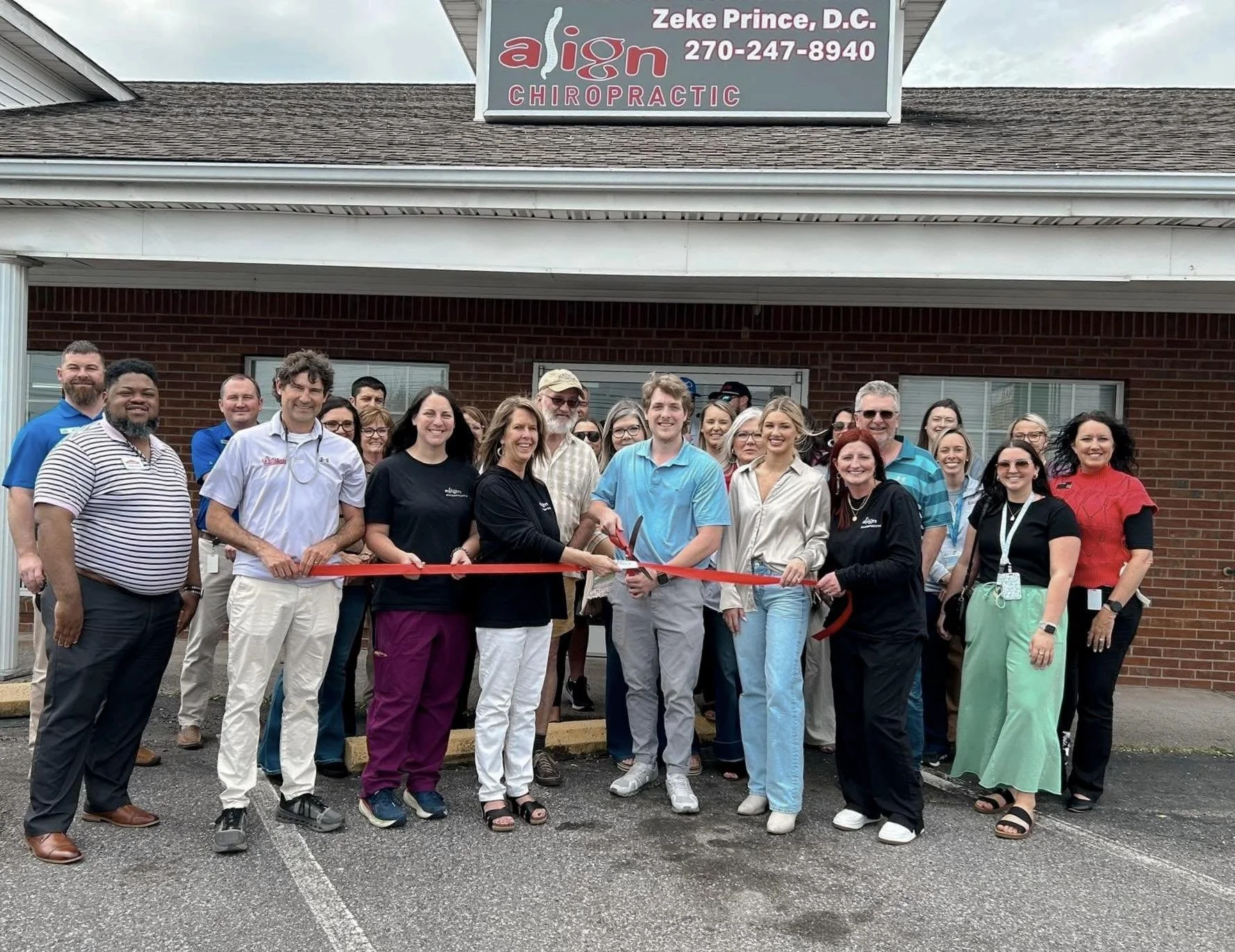 Group of people gathered outside a brick building for a ribbon-cutting ceremony, with some holding the red ribbon and scissors, in front of a sign that reads 'Align Chiropractic'.