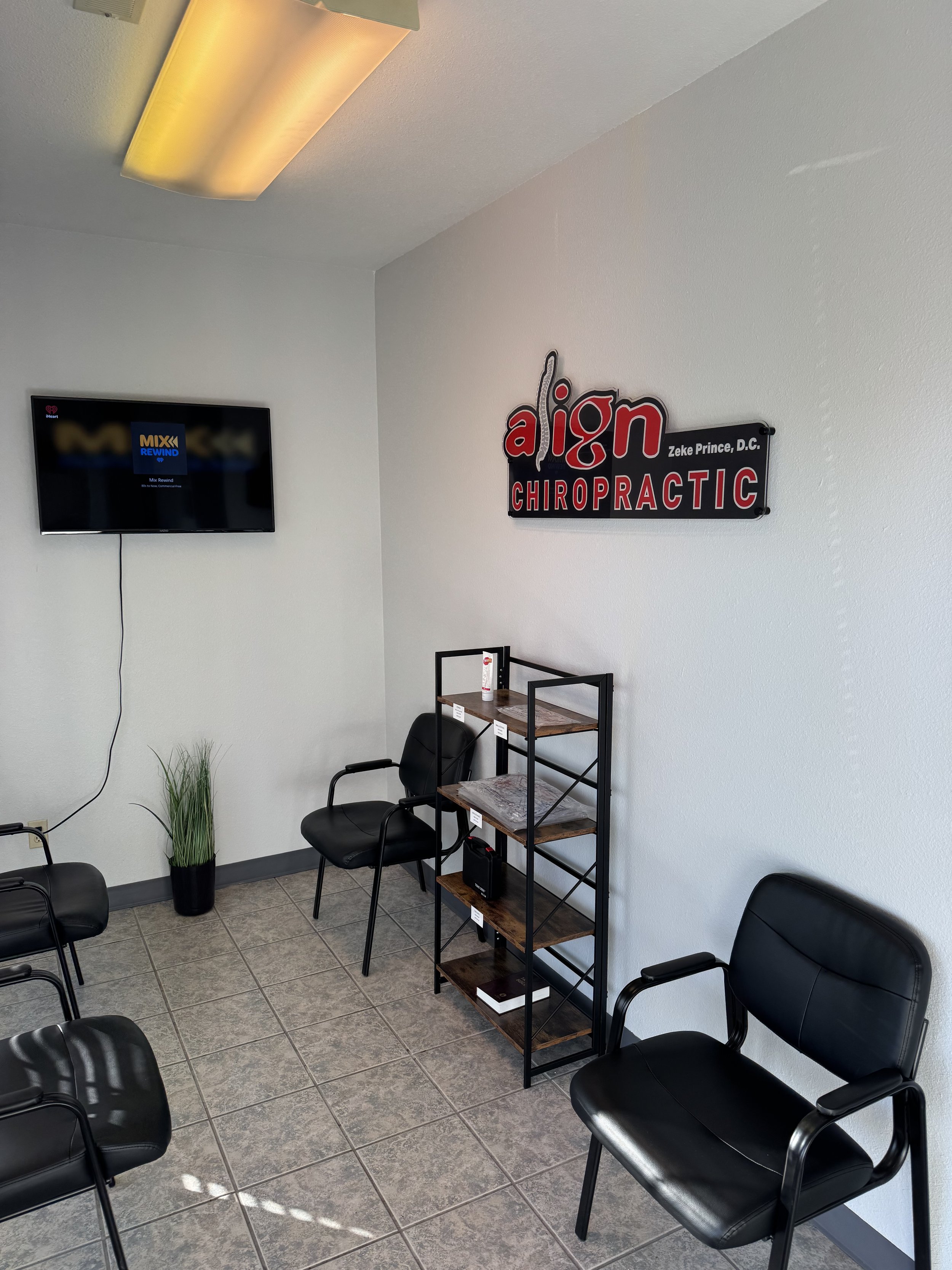 Waiting room at a chiropractor's office with black chairs, a small wooden bookshelf, a potted plant, a wall-mounted TV, and a sign that reads 'align Chiropractic, Zeke Prince, D.C.'