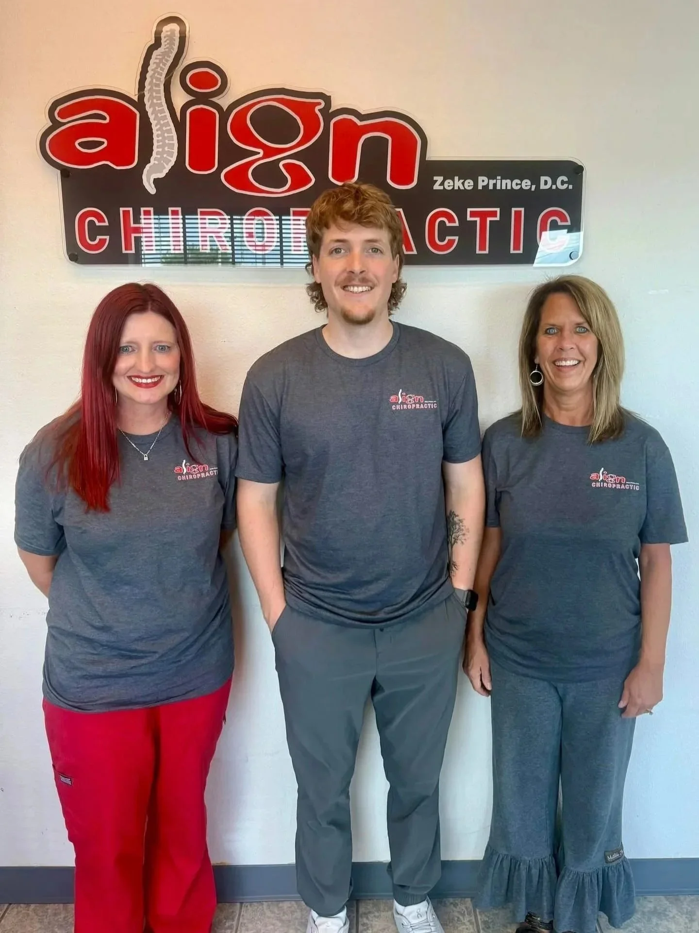 Three people standing in front of a sign for a chiropractic office, all wearing gray shirts with the office logo. The person in the middle has curly hair and tattoos, the woman on the left has red hair, and the woman on the right has blonde hair.