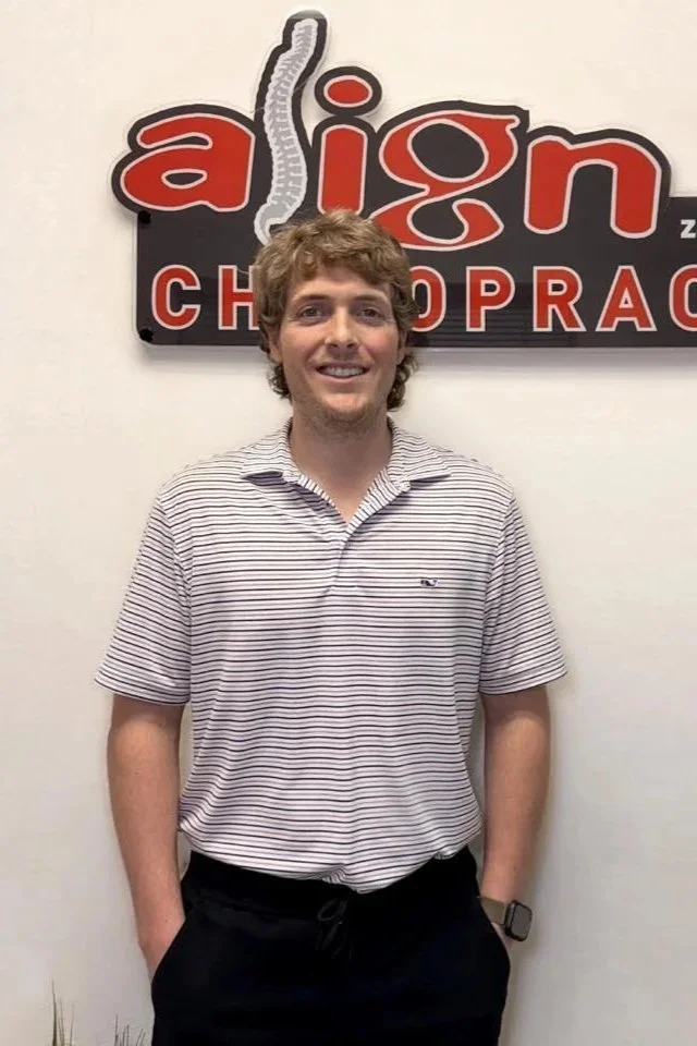 A young man with curly hair and a striped polo shirt stands in front of a sign that says 'align CHIROPRA' with part of the text obscured.