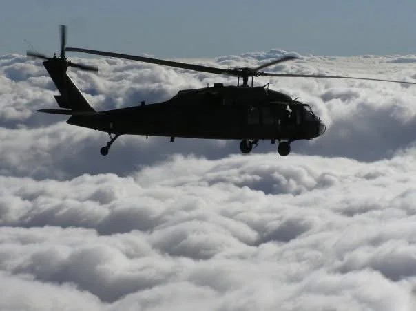 Silhouette of a military helicopter flying above clouds on a clear day.