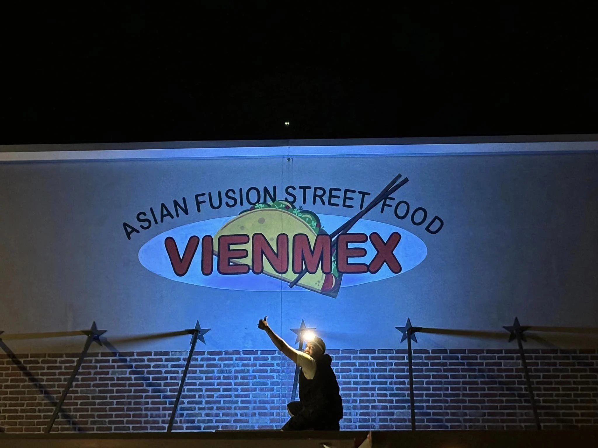 Person sitting in front of a large illuminated sign for Vienmex, an Asian fusion street food restaurant, at night. The sign features a taco with chopsticks, and the person is giving a thumbs-up.