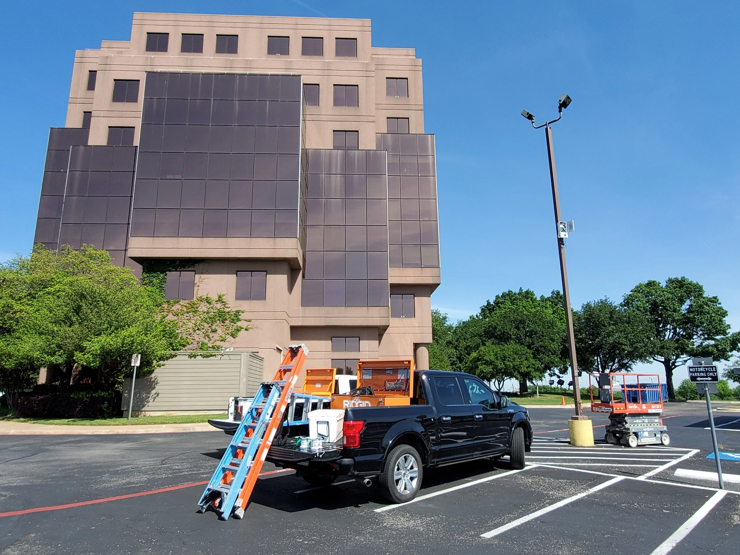 Loading and maintenance equipment in a parking lot near a large office building with dark glass windows. A black pickup truck is parked with a ladder leaning against it, and a cherry picker lift nearby, with a sign indicating motorcycle parking only.