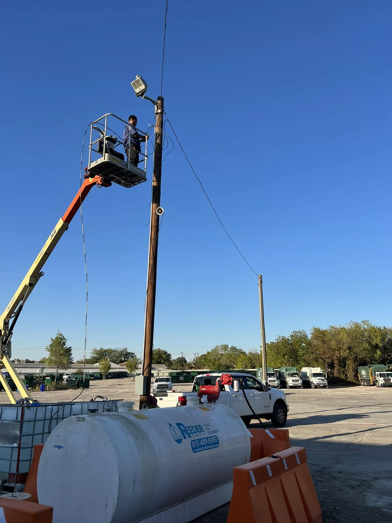 A worker in a lift bucket repairing or inspecting a utility pole with wires, near a truck and a large water tank in a parking lot.