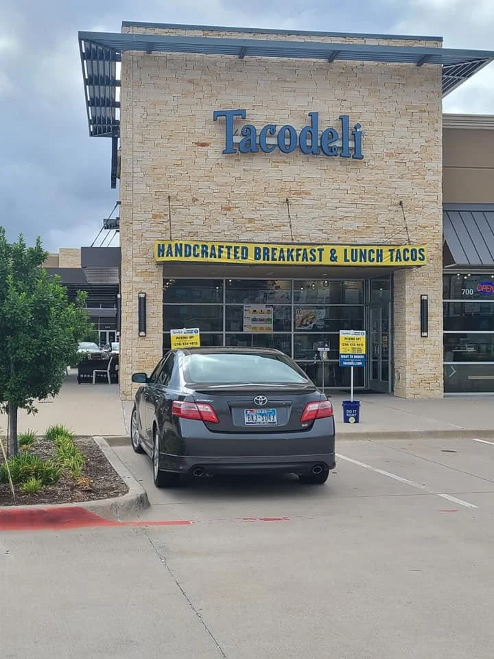 A storefront for Tacodeli, a restaurant that offers handcrafted breakfast and lunch tacos, with a parked black car in front and a small landscaped area to the left.