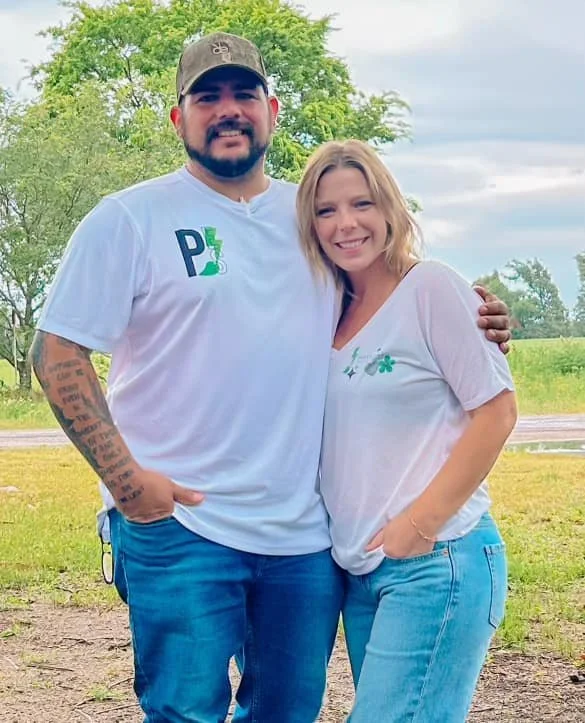A man and woman standing outdoors on a cloudy day, smiling, with greenery and trees in the background. The man is wearing a white t-shirt with a logo, a baseball cap, and has tattoos on his arm. The woman is wearing a white t-shirt with a small green shamrock and light blue jeans.