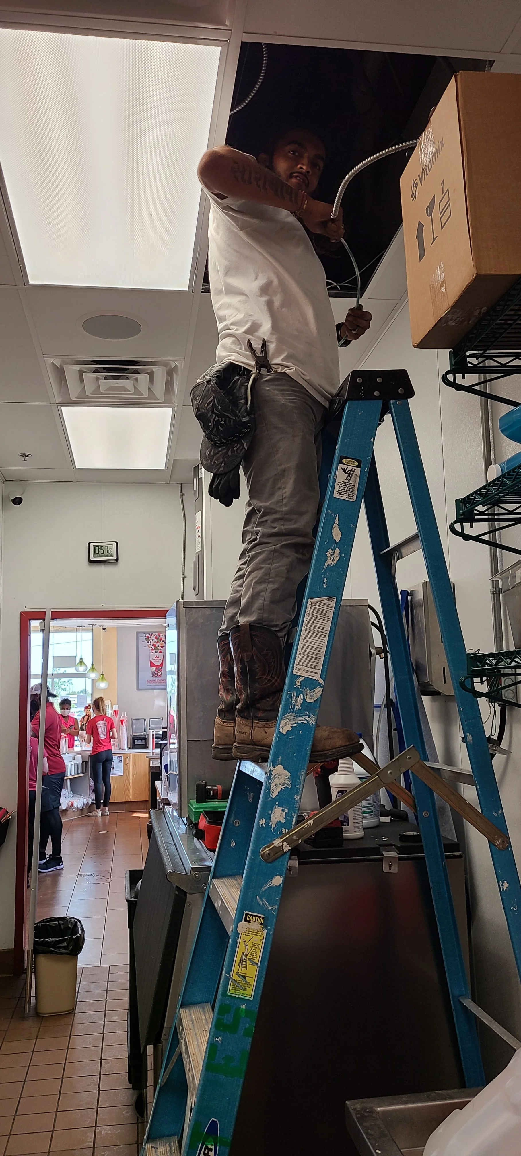 A worker on a blue ladder installing or repairing an ceiling vent or light fixture in a commercial kitchen or restaurant. There are people in pink uniforms working in the background.