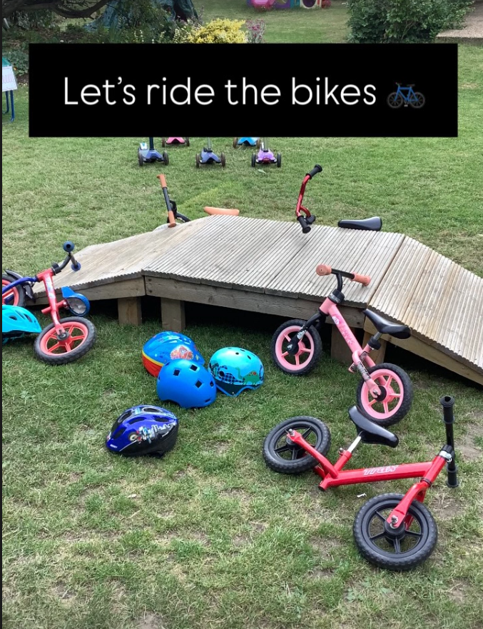 Children's outdoor play area with bikes, helmets, and a small wooden ramp. The sign says 'Let's ride the bikes' with a bicycle icon.