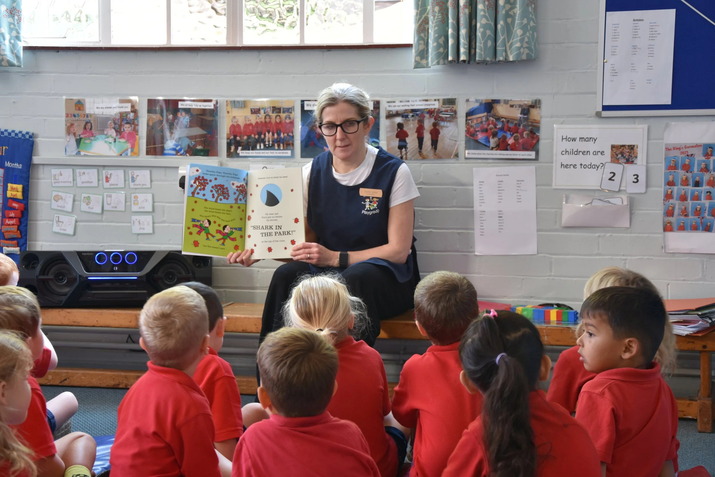 A teacher reading a children's book to a group of young students in a classroom. The students are sitting on the floor, wearing red uniforms, and facing the teacher who is holding the book titled 'Shark in the Park!'