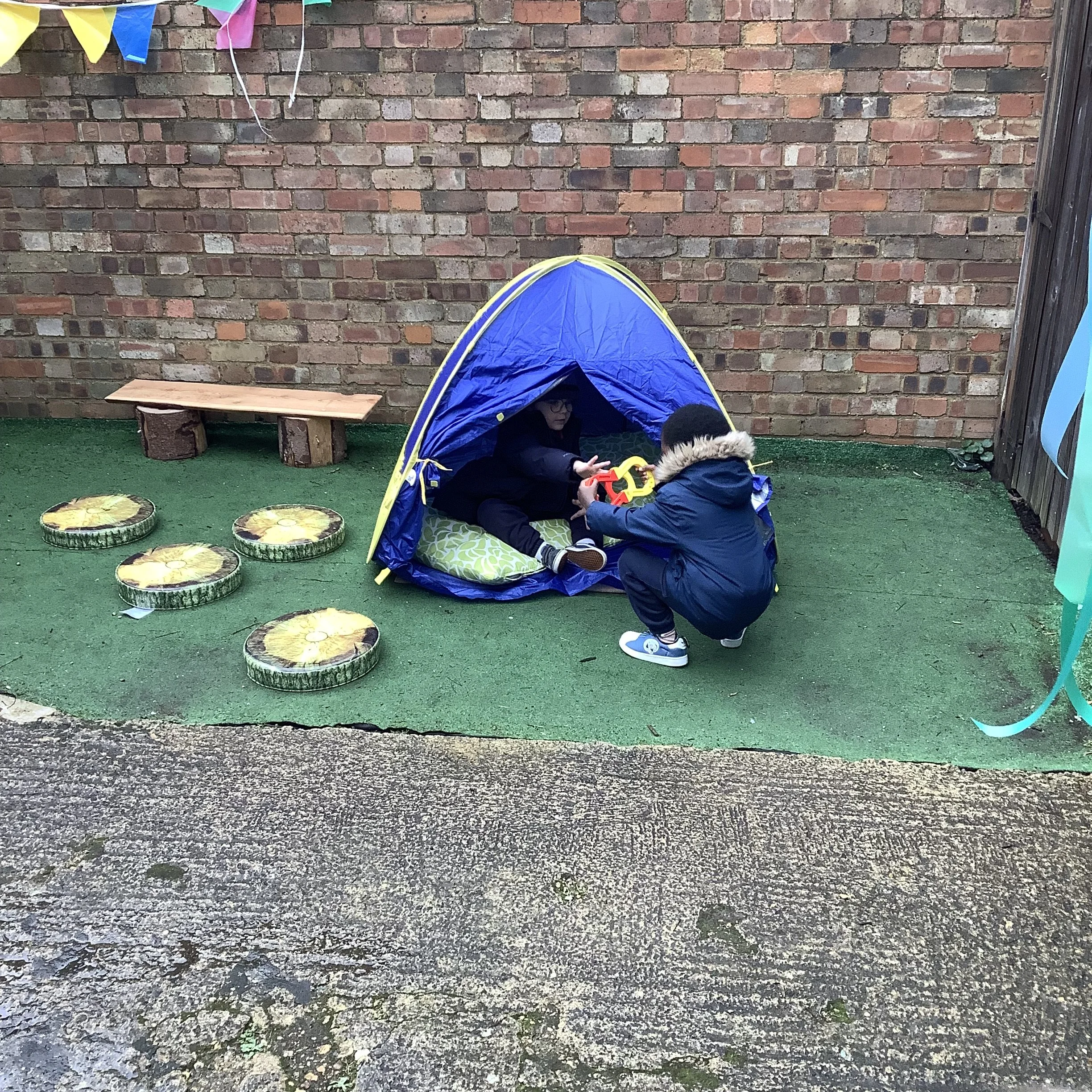 Two children playing outside with a small blue tent, one sitting inside and the other handing an object to the seated child, with decorative watermelon-shaped cushions, a small wooden bench, and a brick wall in the background.