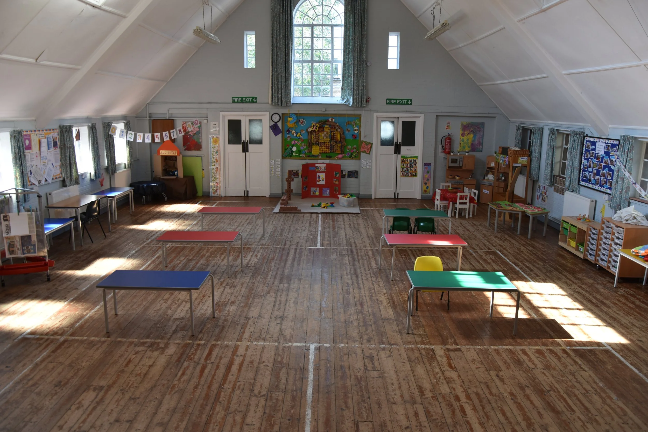 Empty kindergarten classroom with decorated walls, small tables and chairs in various colors, and play areas with toys and books.