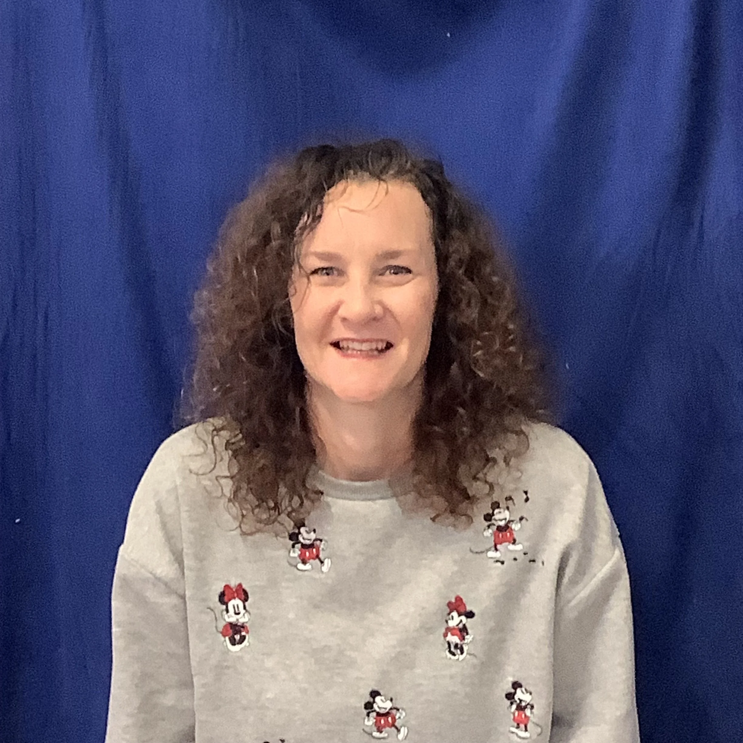 A woman with curly hair smiling, wearing a sweater with printed images of Minnie Mouse, standing in front of a dark blue curtain.