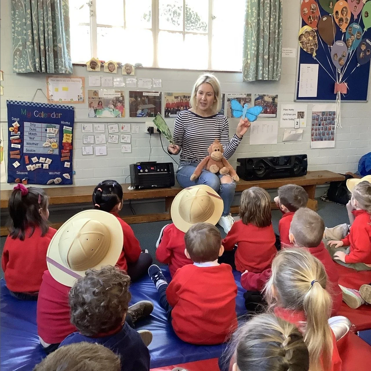 A woman sitting on a desk reading a story to a group of children sitting on mats. She is holding a blue butterfly puppet in one hand and a green book in the other. The children are in red uniforms and some are wearing hats. The classroom has educational posters on the walls, including a calendar and photos of children.
