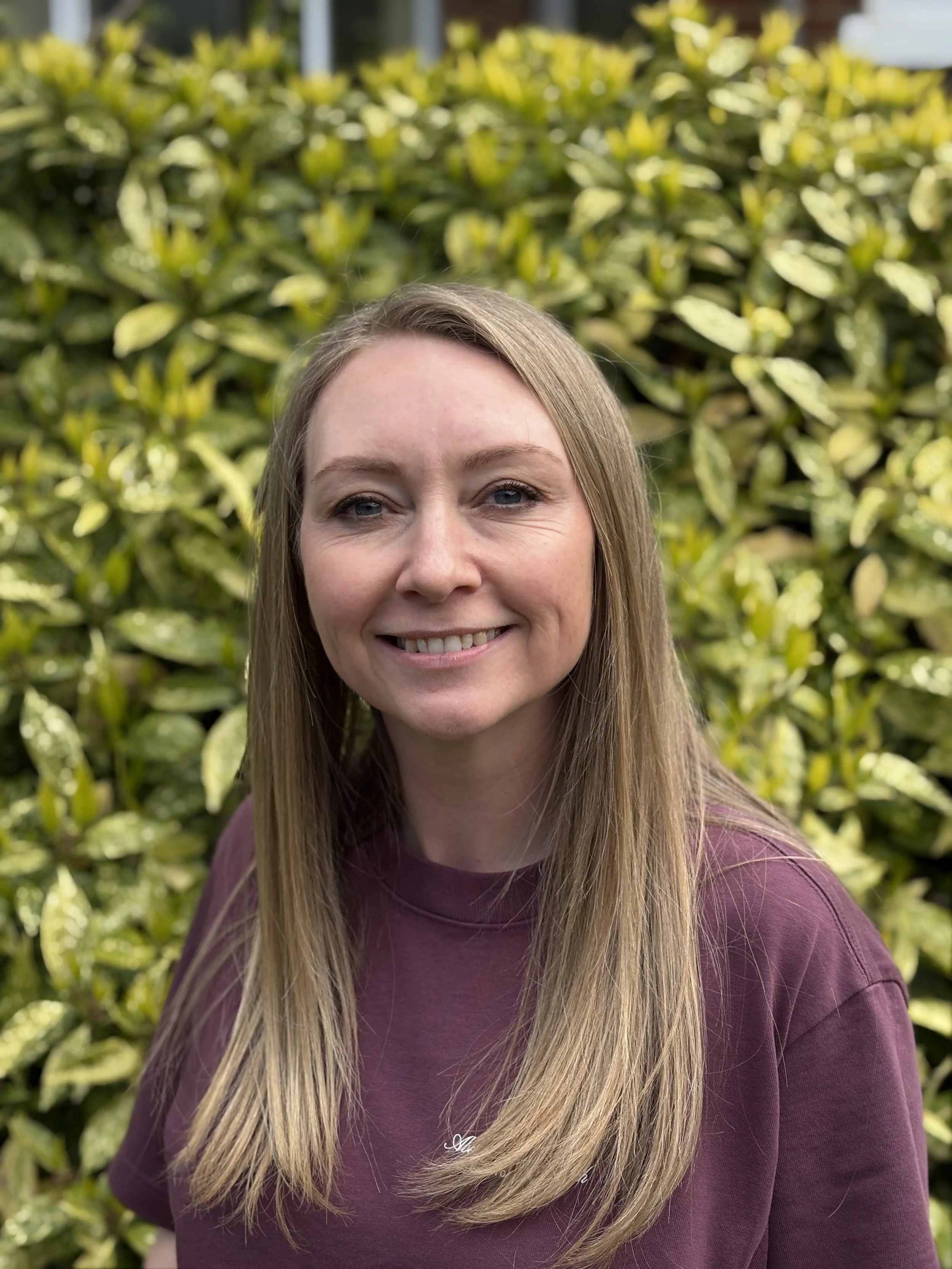 A woman with long blonde hair and blue eyes smiling outdoors with green bushes in the background.