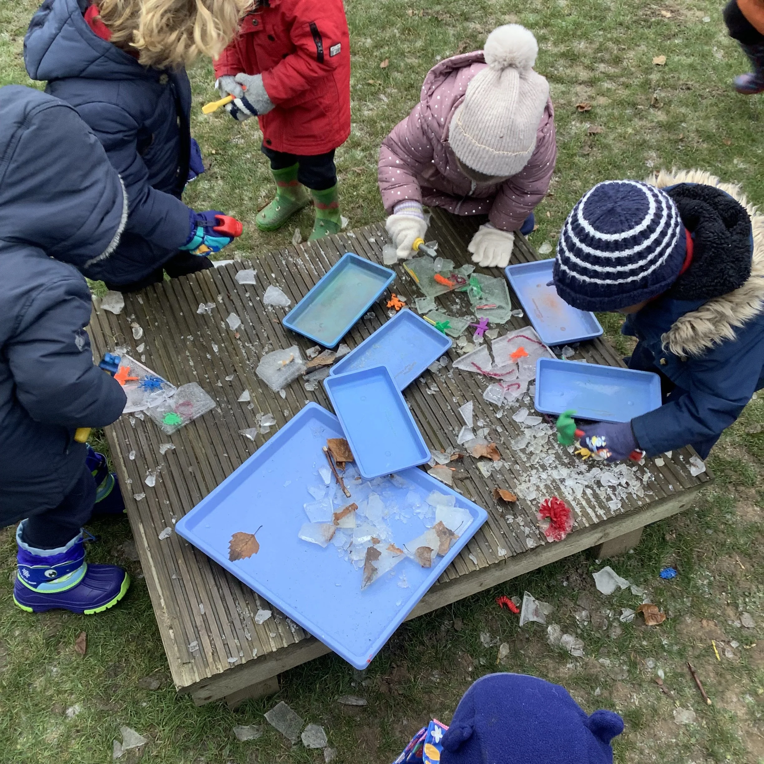 Children playing outdoors with broken glass and toys on a wooden table, wearing winter clothing and gloves.