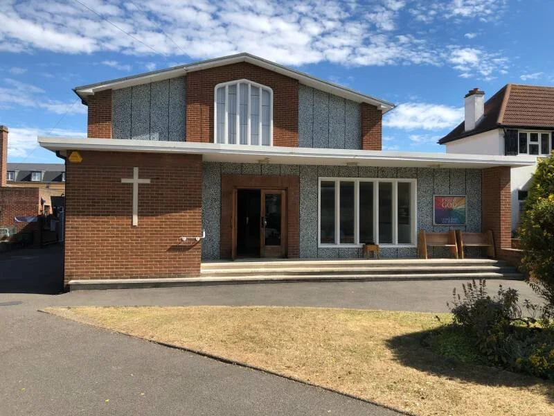 A church building with a cross on the brick wall, large front windows, and wooden benches outside under a flat roof, with a grassy area and pathway in front.