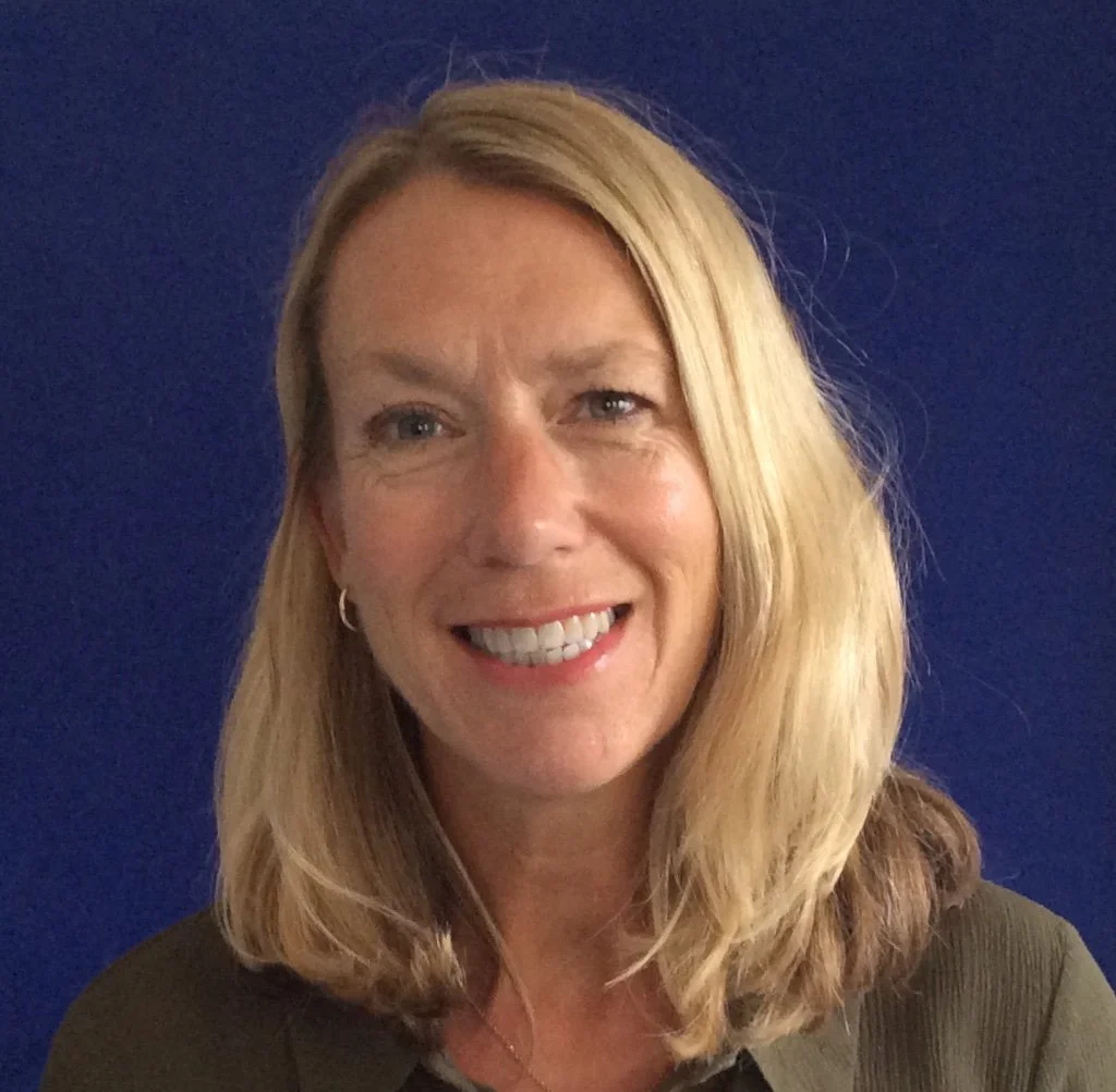 A woman with shoulder-length blonde hair smiling in front of a dark blue background.