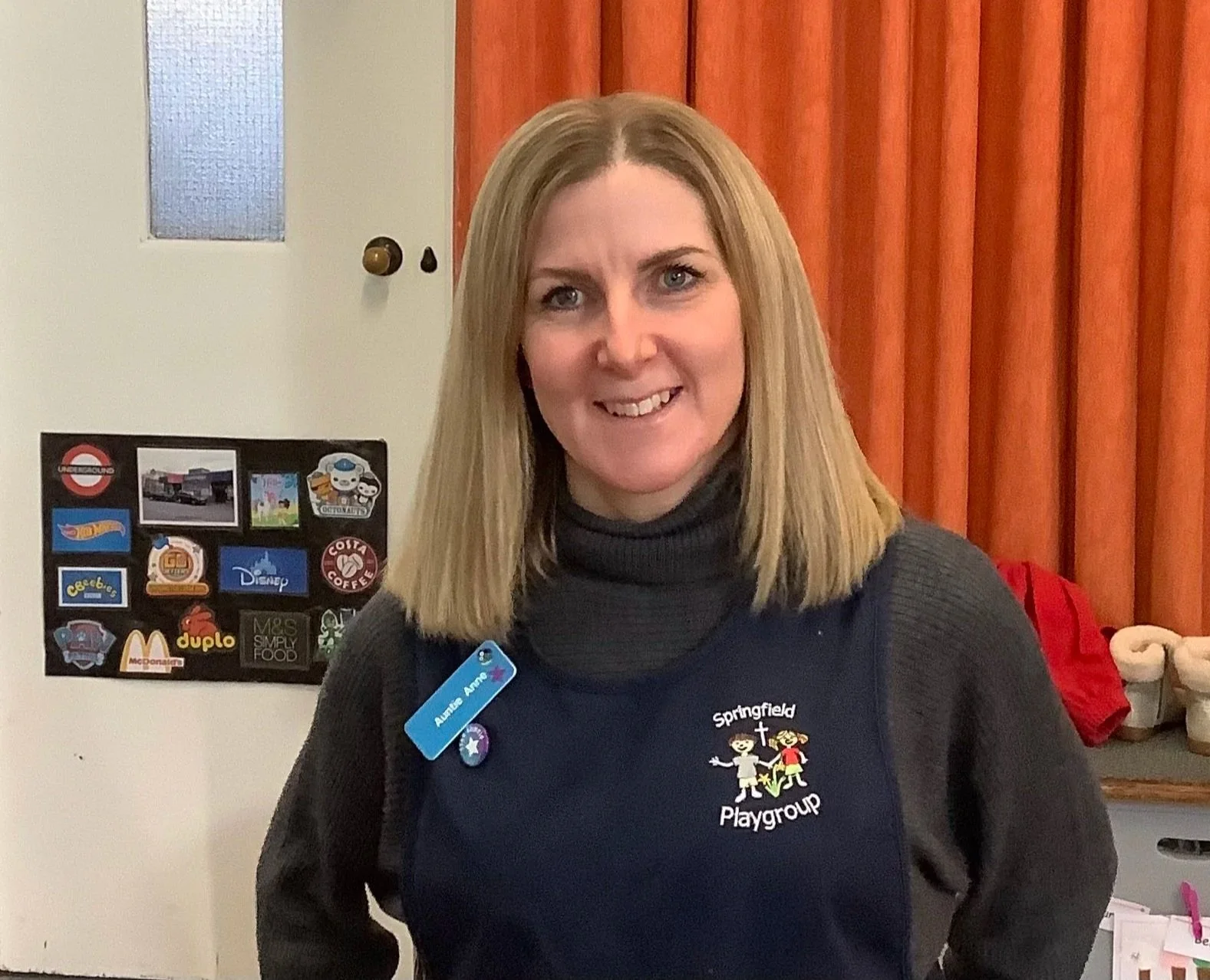 A woman with shoulder-length blonde hair smiling at the camera, wearing a black Springfield Playgroup shirt with a name tag that reads 'Austine Anne', stands indoors in front of a white wall with colorful stickers and a red curtain.