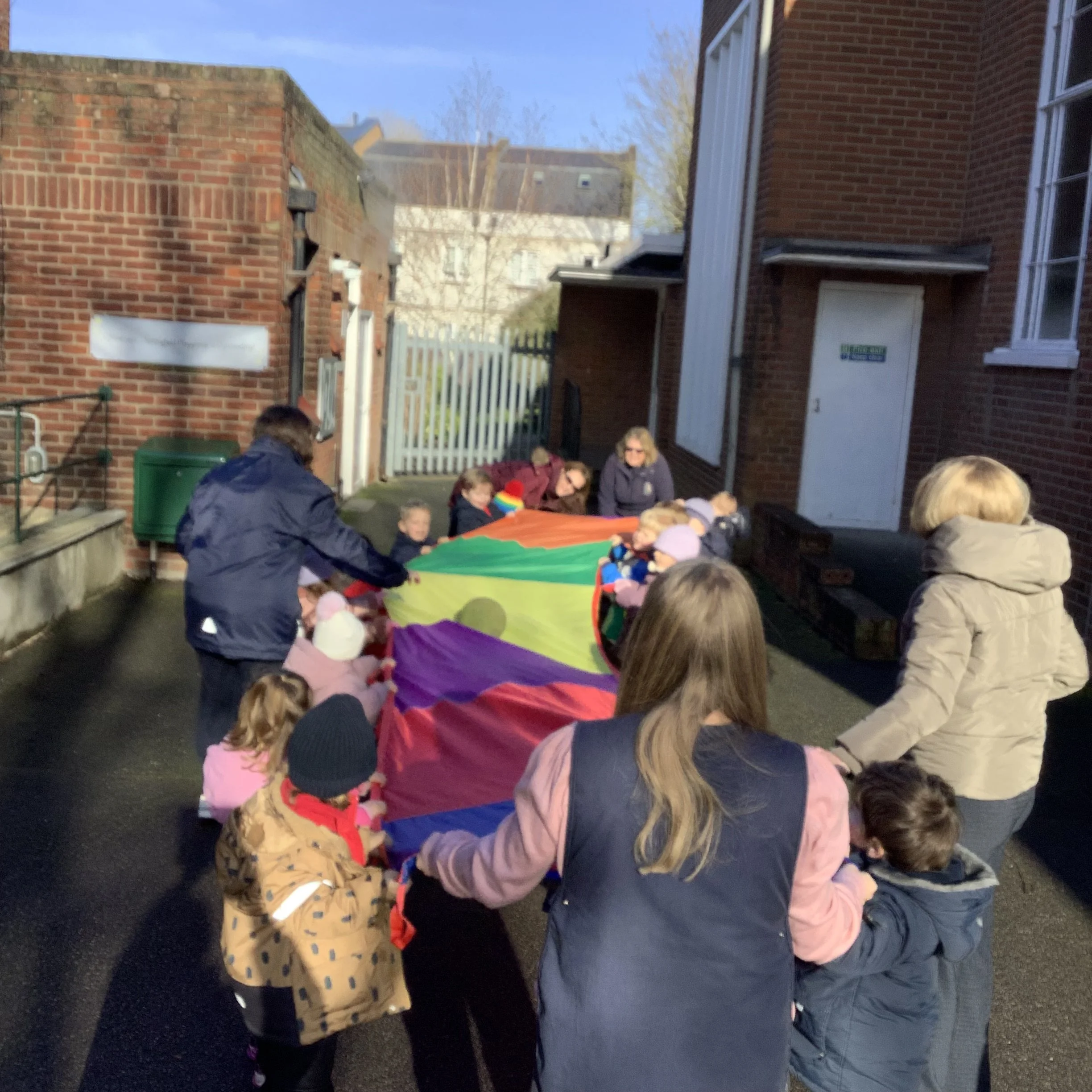 Group of children and adults holding a colorful parachute outdoors in a small yard, with brick buildings and a white gate in the background.